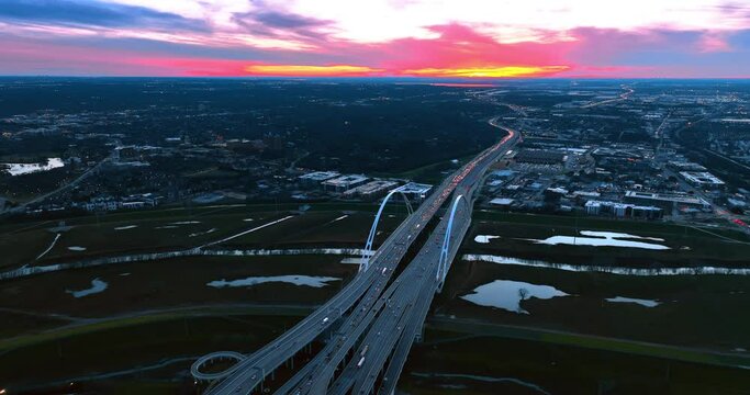 Multiple cars with headlights on move by the Dallas Margaret Mcdermott Bridge at twilight time. Spectacular view of the metropolis from top with colorful sky at backdrop.