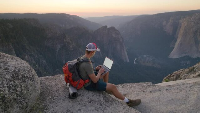 A hiker sits on a mountain edge with a laptop, taking in the amazing view of Yosemite Valley as the sunset paints the sky. Slow Motion, Camera 4K RAW. 