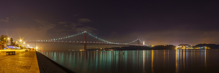 Panorama of illuminated red bridge 25 de Abril Bridge crossing the Tagus river with statue of Cristo Rei during night time, Lisbon, Portugal