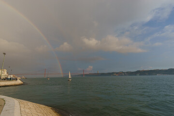 Rainbow over red bridge 25 de Abril Bridge with statue of Cristo Rei sailing boat during sunset, Lisbon, Portugal