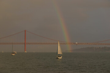Rainbow over red bridge 25 de Abril Bridge with sailing boat during sunset, Lisbon, Portugal