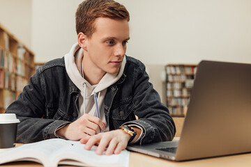 Male student in a library reading from a laptop. University student with ginger hair sitting at a desk.