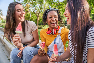 Group of young multiracial female companions laughing enjoying ice cream in park on summer day. Three funny women smile excitedly while chatting outdoors. Generation z and friendship in youth. - obrazy, fototapety, plakaty