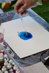 A gloved hand performing alcohol ink art by applying vibrant blue alcohol ink with a pipette on white paper, showcasing a vivid fluid pattern