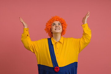 a cheerful girl in a clown costume and a bright wig raised her hands up on a colored background