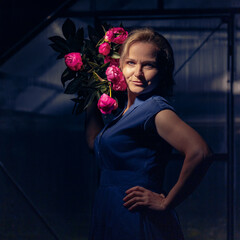 Confident woman holding pink peonies in a greenhouse, with dramatic lighting