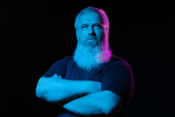 Man with a serious expression, crossed arms, and a vivid pink beard under blue lighting against a...