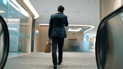 Back view of skilled business man phone calling to project manager while walking to meeting room. Caucasian project manager walking with meeting room with blurred background. Full body. Exultant.