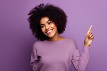 Radiant Young Woman with Curly Afro Hair Pointing Upwards on a Lavender Background. Lifestyle and...
