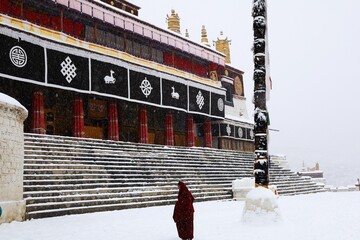 Amidst a serene blanket of snow, a solitary Tibetan monk clad in maroon robes gracefully paces...