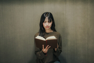 A young Asian woman is sitting by the window in a cafe reading a book.
