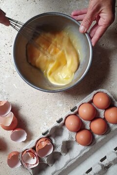 directly above shot of person whisking eggs in stainless steel bowl with fresh eggs in carton on stone table