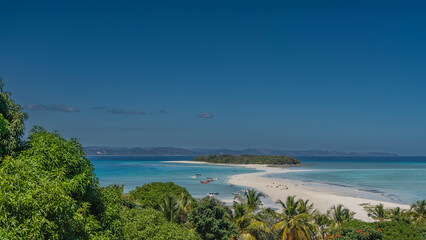 A beautiful tropical island in the aquamarine ocean. Tiny silhouettes of people are visible on the sandy spit of the beach. The boats are moored. Green vegetation in the foreground. Nosy Iranja  