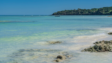 The beautiful aquamarine ocean stretches to the horizon. The waves are foaming on the coastal rocks. The boat is anchored in the distance. A green hill against a clear blue sky. Madagascar.