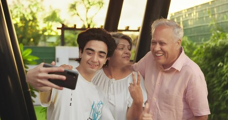Indian Asian happy old aged mom dad couple pair teen child young adult boy hold mobile use phone look cam take funny face selfie make fun older elder male female lad family enjoy joy home park garden