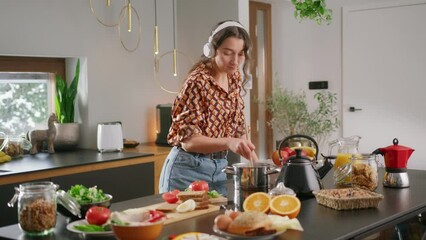 Girl wearing headphones dances in kitchen while stirring pot with wooden spoon