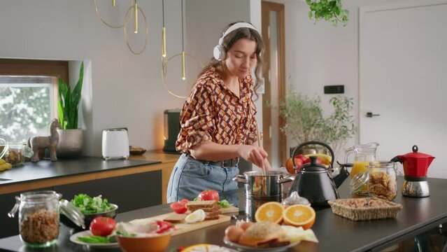 Girl wearing headphones dances in kitchen while stirring pot with wooden spoon