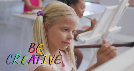 Fototapeta premium Image of be creative text in multi colours over happy caucasian schoolgirl painting in art class