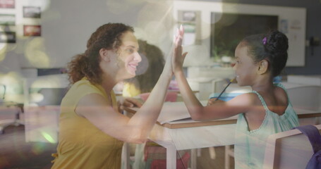Image of bokeh lights over diverse female teacher and pupil high fiving at desk in class