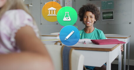 Image of school icons over smiling biracial schoolboy sitting at desk in class