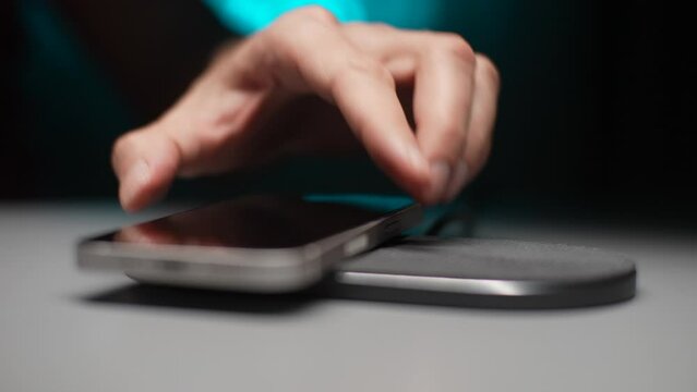Close-up cropped shot of unrecognizable man putting mobile phone on wireless charging pad on desk. Closeup of male charging dead phone with modern wireless charger at home in dark room, slow motion.