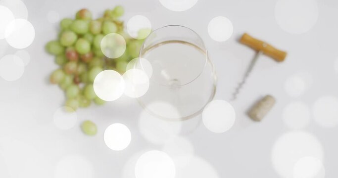 Composite of glass of white wine, corkscrew and white grapes over spots of light on white background