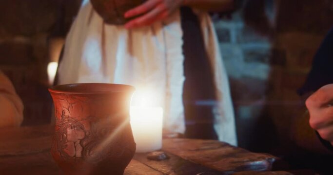 Woman servant pours wine into guest clay mug in tavern. Medieval maid in white apron fills client cup with alcohol in pub closeup. Dark ages inn service