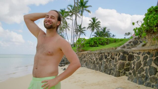 Self-confident happy man with fair hair gathered in a bun standing on a sand beech with palm trees and green lawns over blue sky with fluffy white clouds. High quality 4k footage