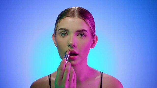 Caucasian young blonde girl paints her lips looking at camera, close up shot in beauty studio
