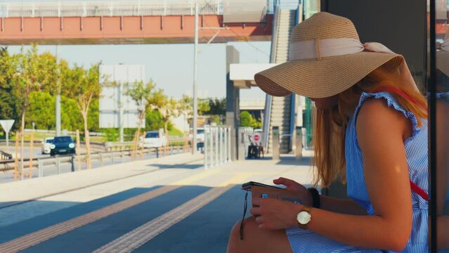 Caucasian white woman sits on a train station and patiently waits for the next train. While waiting she scrolls on her phone.