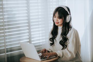 Smiling Asian woman at coffee shop with headphones listening to music and using laptop