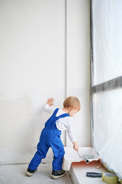 Little child construction worker standing by the window and painting white wall in apartment. Kid in work overalls using paint brush while playing at home under renovation.
