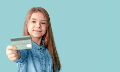 Fototapeta premium A girl in a denim shirt holds a credit card in her hands isolated on a blue background. Copy space
