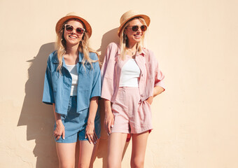 Two young beautiful smiling hipster female in trendy summer clothes. Sexy carefree women posing on street background. Positive models having fun, hugging at sunset. In hat and sunglasses. Near wall