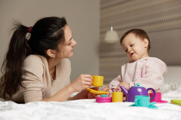 Obraz na płótnie Canvas happy mother and little child daughter pretending drinking tea from toy cups and spending time together in bedroom, family having fun