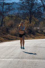 A young happy runner girl, dressed in running shorts and a tight top, running suspended in the air with style and perfect technical movements on an asphalt road casting her shadow on the asphalt.
