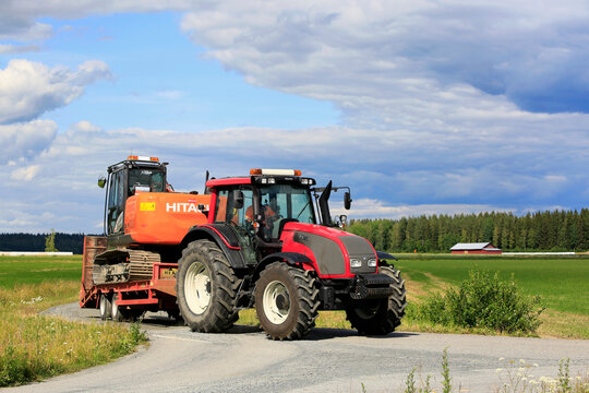 Red Valtra T151 farm tractor transports Hitachi Zaxis 130 LCN excavator along country road on sunny day of summer.