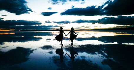 young two woman dance across the mirror like surface of salt lake as the sun disappears behind the...