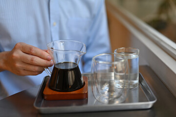 Cropped with a male's hand holding a small glass pitcher with black coffee on the tray in the cafe