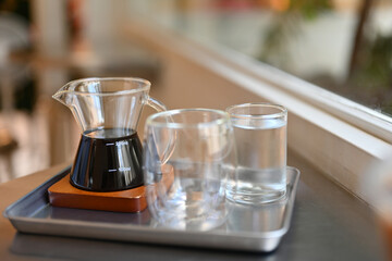 Freshly brewed coffee in a small glass pitcher with an empty and water glasses on the tray in the...