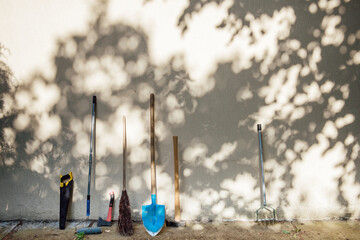Tools for the care of the garden and the house stand near the gray wall in the shade of trees and the rays of the sun. A real life shot with space for text, banner size - obrazy, fototapety, plakaty