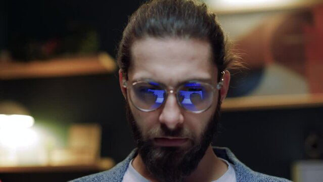 Concentrated seriously looking young man typing using laptop sitting on couch in his living room. Male employee in glasses working remotely on computer, writing report at night time.