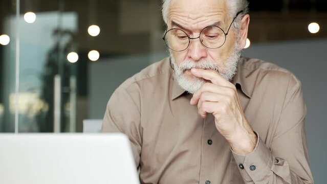 Thoughtful senior gray haired businessman thinking about problem solving while working on laptop sitting at workplace in business office. Elderly professor in glasses is busy with project on computer