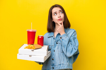 Young Ukrainian woman holding fast food isolated on yellow background having doubts