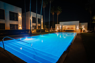 Fototapeta na wymiar Sotogrante, Spain - January 27, 2024 - Night view of an illuminated blue pool with surrounding palm trees and a modern building with lights.