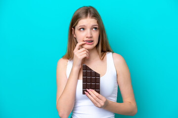 Teenager Russian girl isolated on blue background taking a chocolate tablet and having doubts