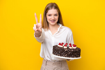 Teenager Russian girl holding birthday cake isolated on yellow background smiling and showing...