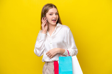 Teenager Russian girl isolated on yellow background holding shopping bags and calling a friend with...
