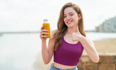 Young redhead girl holding an orange juice at outdoors with thumbs up because something good has happened