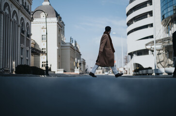 A fashionably dressed individual walks with purpose through a cityscape, exuding confidence and style against an urban backdrop.